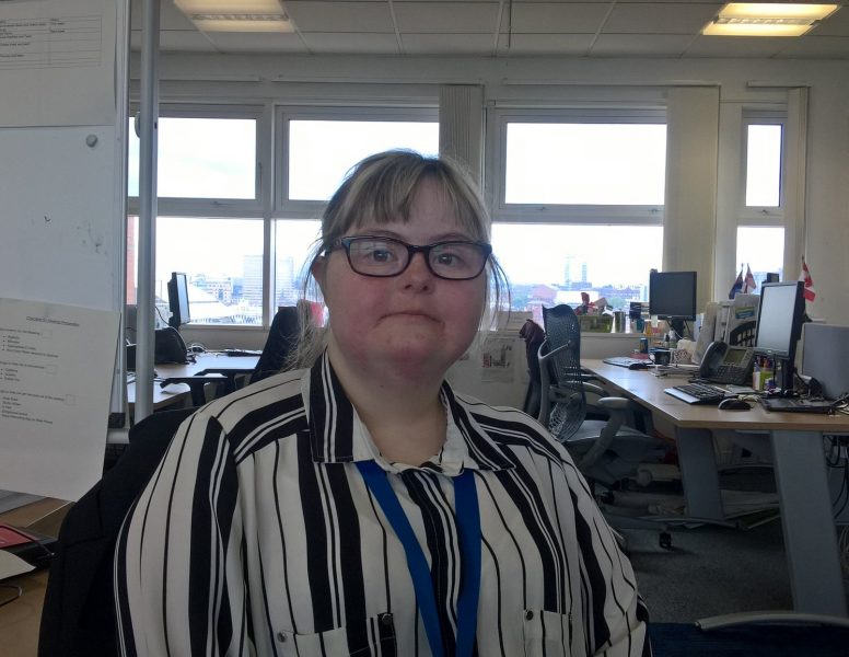 Katie in her office wearing a stripy shirt, looking at the camera with desks and windows behind