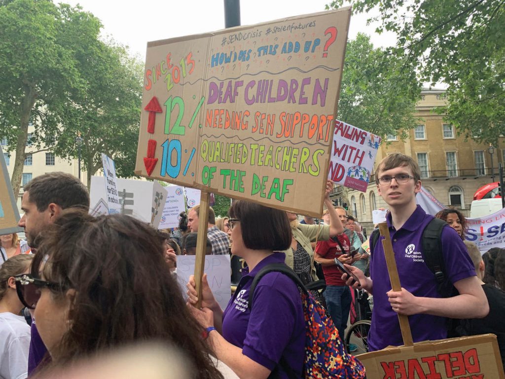 Crowd with woman holding placard saying "How does this add up? Since 2015 12% increase in deaf children needing support, 10% decrease in qualified teachers of the deaf"