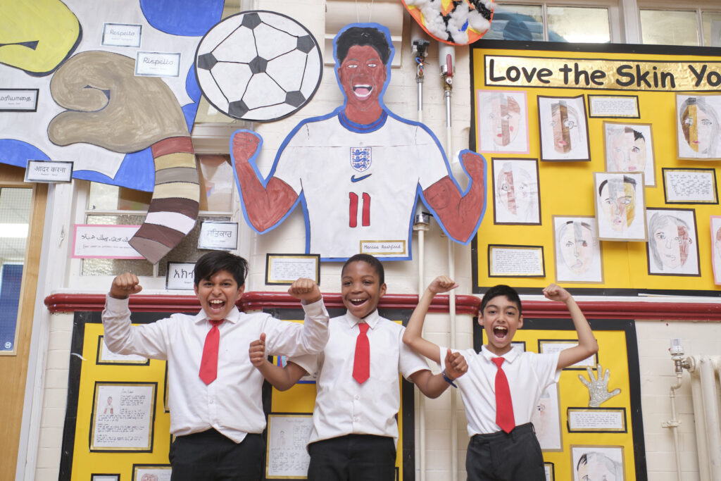 Photograph of pupils at Monega Primary School in Newham. Three boys in school uniform make celebration poses in front of a collage with footballer Marcus Rashford in a similar celebratory pose. Image copyright: Monega Primary School.