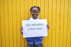 Monega Primary School 5, pupil holding a sign with the text 'Our education is fisrt class!'. Copyright: Monega Primary School