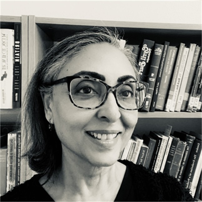 A black and white photograph of a woman smiling. She is wearing large tortoise-shell glasses, a black shirt and earrings. She has dark shoulder length hair and is smiling broadly. Behind her is a bookshelf full of new and old academic books.