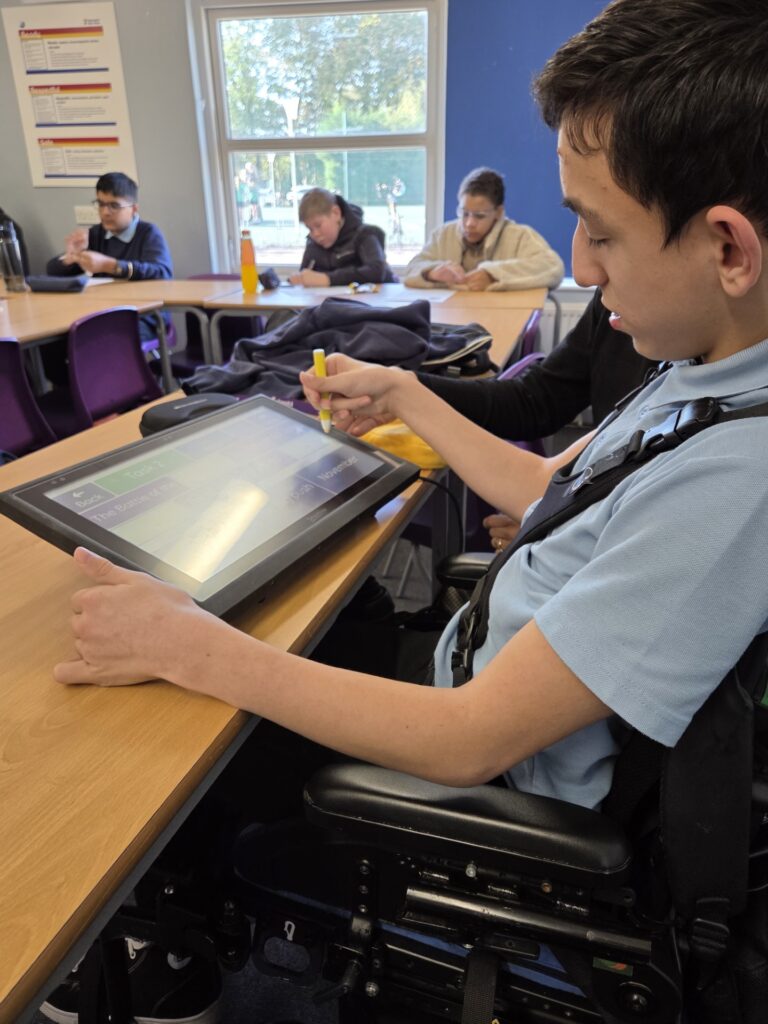 A secondary school student in a classroom using a screen and stylus to complete a task. He has short brown hair and is sitting at a desk in a black wheelchair. His face is in profile as he focusses on the screen, moving the stylus with the support of an unseen assistant. There are three other students in the classroom working independently, with a window behind them.