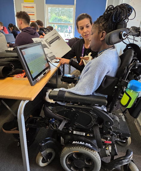 A secondary school student working in a classroom using a screen and booklet supported by a teaching assistant. The student is wearing a pale blue jumper and her long black braided hair is tied up in a high bun. She is sat at her desk in a motorised wheelchair focusing on a screen showing a keyboard. Next to her a woman holds a printed booklet and is pointing to the text. The classroom is full of other students working.
